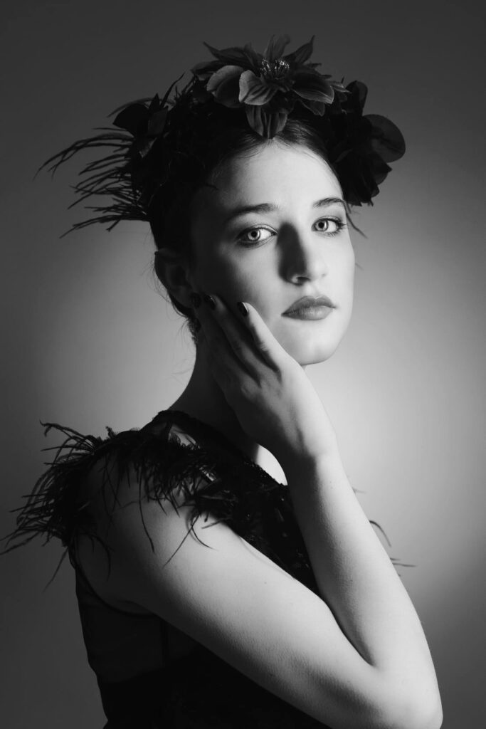 Black and white portrait of a young woman with her hand touching her cheek, gazing intently towards the viewer. She wears a dark textured dress and a headpiece adorned with dark leaves and flowers, adding a dramatic and artistic touch to her appearance.