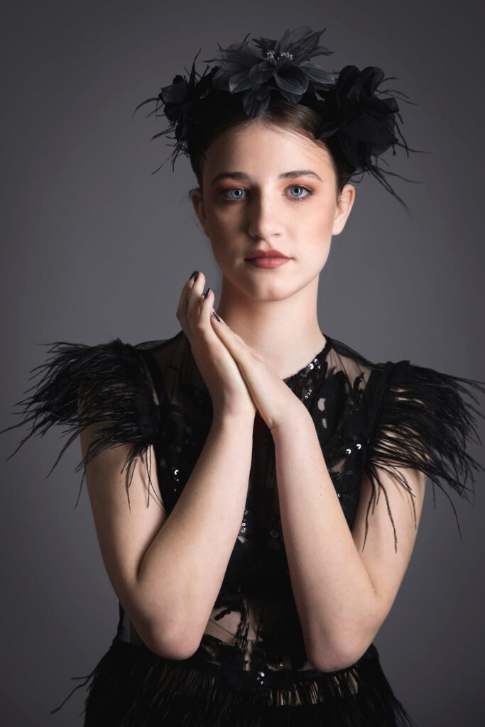 Portrait of a young woman with pale skin and dark hair styled in an elegant updo adorned with a black floral headpiece, captured by a Cotswolds photographer in Gloucestershire. She is wearing a black dress with feathered shoulders and sequin details, holding her hands gracefully near her chin against a soft grey background