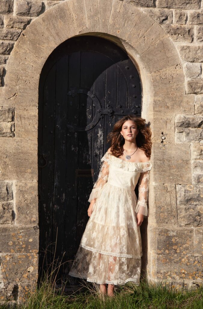 Young woman with flowing brunette hair standing in front of an old arched black door set in a stone wall. She wears an elegant, off-the-shoulder cream lace dress and looks slightly away from the camera, with sunlight highlighting her hair and dress